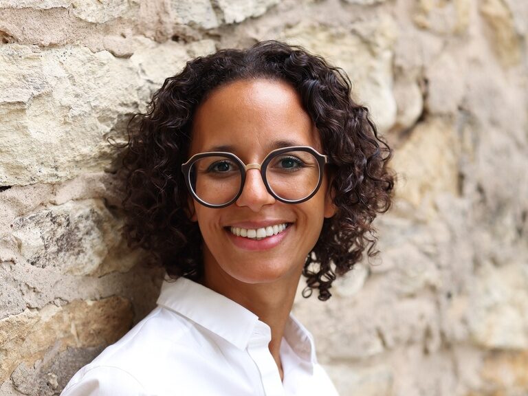 lächelnde Wellnesscoach Claudia Schober mit Brille vor einer Steinwand.