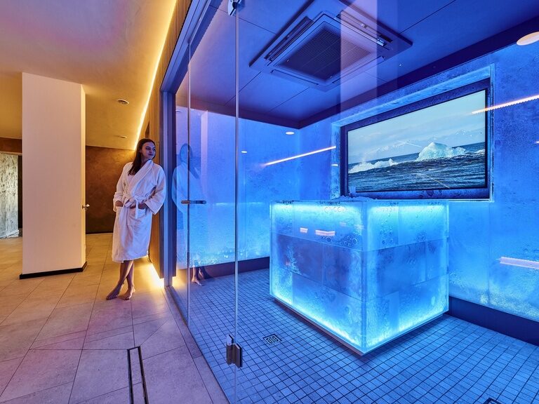 Eine Frau in Bademantel lehnt entspannt an dem prächtig beleuchteten Eisbrunnen im Wellnessbereich des Hotels der Birkenhof in der Oberpfalz.