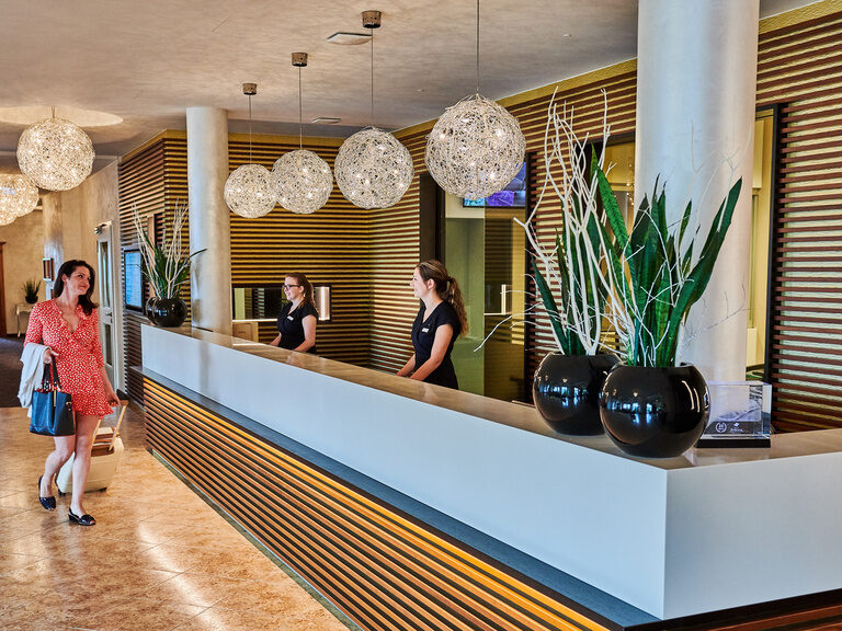 Die freundlichen Rezeptionistinnen im Hotel Der Birkenhof empfangen eine Frau im roten Kleid.