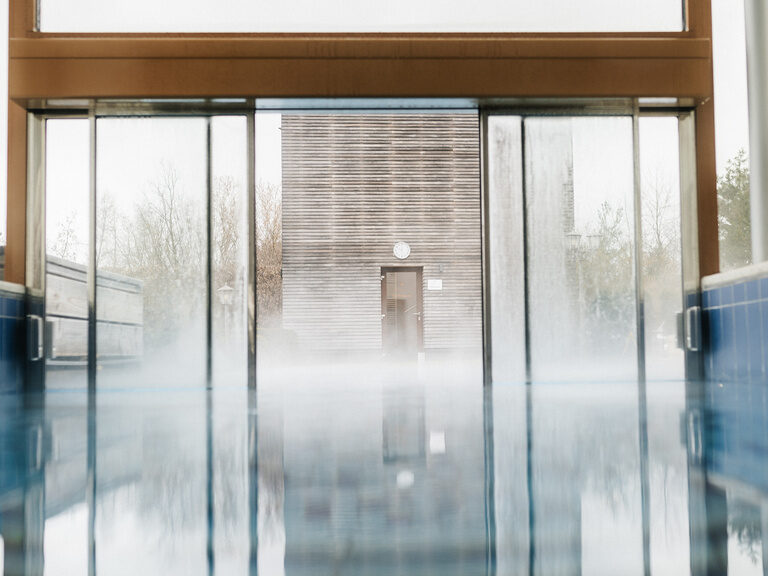 Der modern gestaltete Indoorpool des Wellnesshotels der Birkenhof in Bayern mit der Durchgangstür zum Aussenbereich der Wasserwelt.