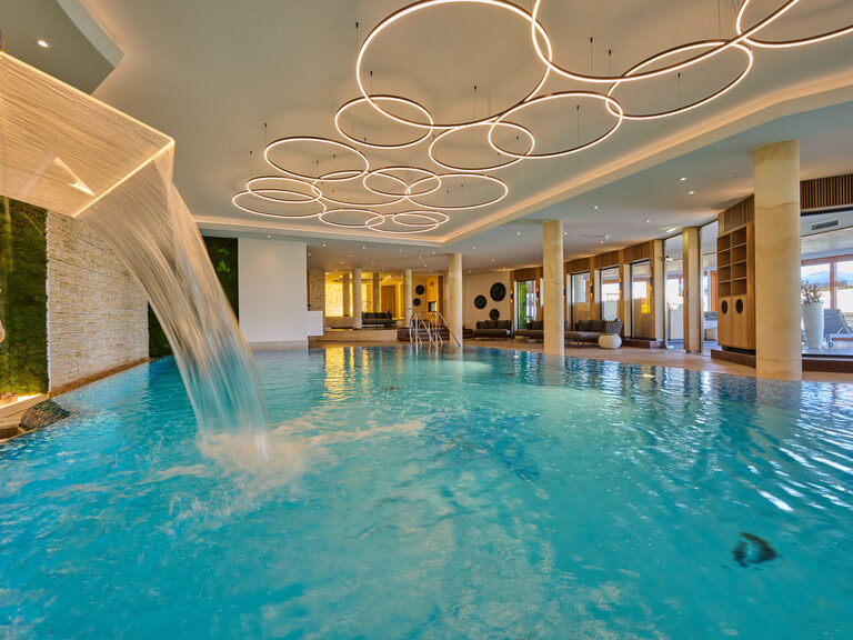 Das elegante Hallenbad mit einem zentralen Wasserfall im Wellnesshotel Der Birkenhof in der Oberpfalz.