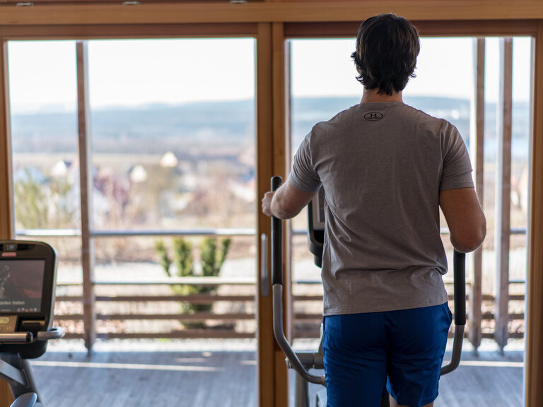 Ein Mann trainiert im Sportbereich des Hotels der Birkenhof in der Oberpfalz mit Panoramaausblick, umgeben von moderner Fitnessausstattung.
