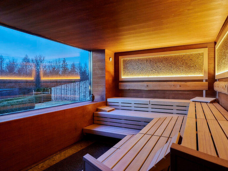 Holzbänke in der Panoramasauna vor einem Fenster mit Blick in den Wellnessbereich des Hotels der Birkenhof beim bayerischen Wald.