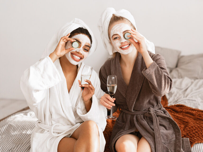 Zwei Frauen mit Gesichtsmaske, Gurkenscheiben und Sektgläsern genießen den Tag im Bademantel.
