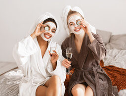 Zwei Frauen mit Gesichtsmaske, Gurkenscheiben und Sektgläsern genießen den Tag im Bademantel.
